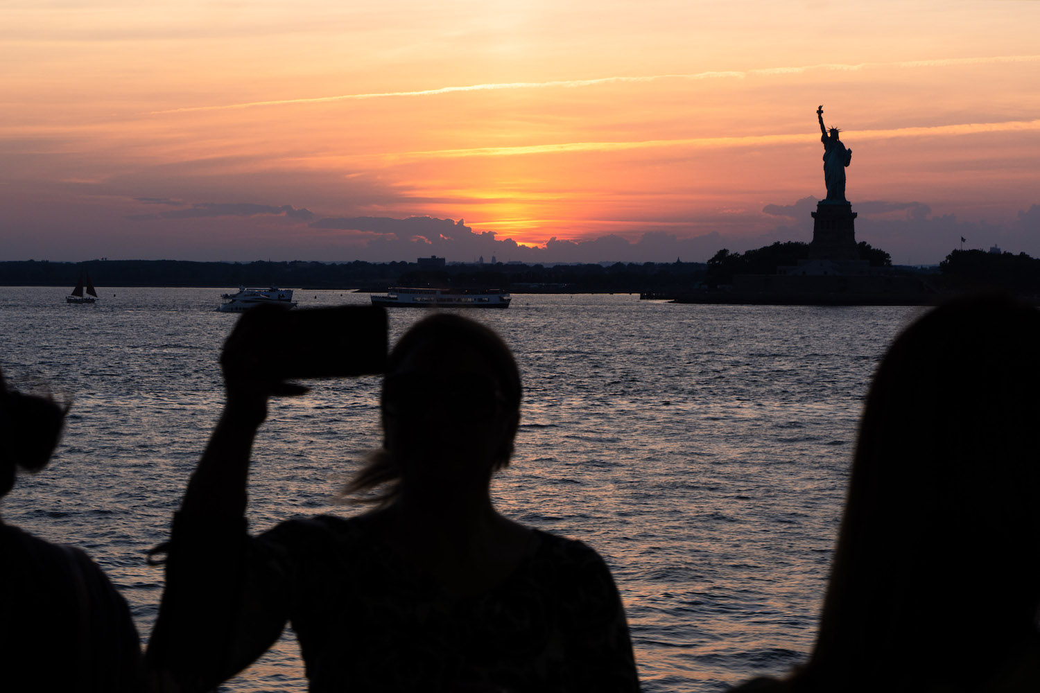 The silhouette of three people looking at the Statue of Liberty onboard the Staten Island Ferry. The sun is setting below the horizon.