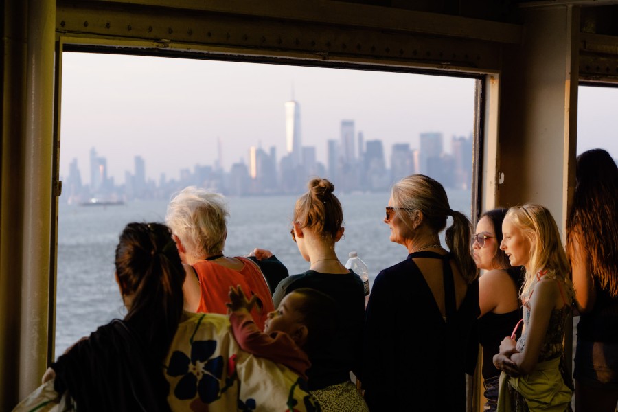 Several people stand by the window on the Staten Island Ferry with the sun shining on them. In the background is the downtown Manhattan skyline.