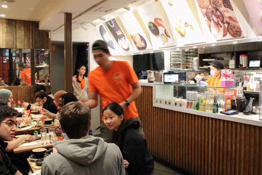 In the left foreground of the image, three customers are sitting at a table in the sports bar. A waiter dressed in an orange shirt is captured in motion and blurry next to them.