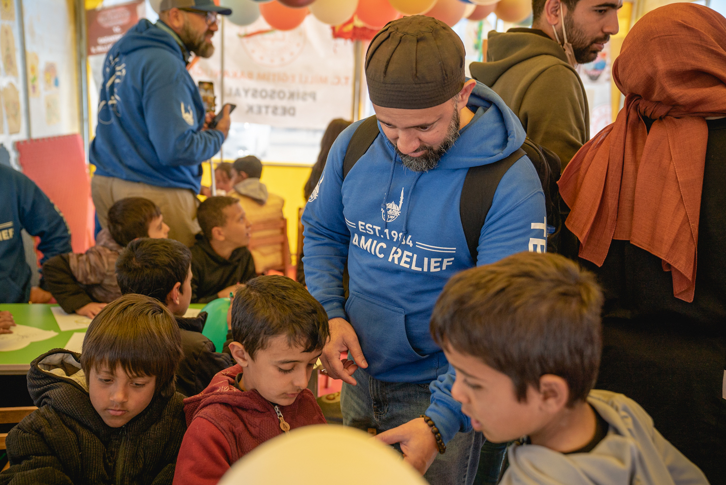 A person wearing a blue hoodie with white text “E.S.T. nineteen eighty-four” and “Islamic Relief” on the front stands among a group of children.