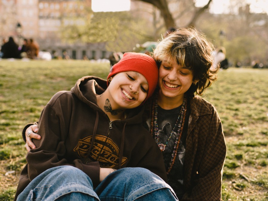 Koda Fraga and Leo Koulish sit together on the grass in Washington Square Park with the sun shining on them. They are holding each other and smiling at the camera.