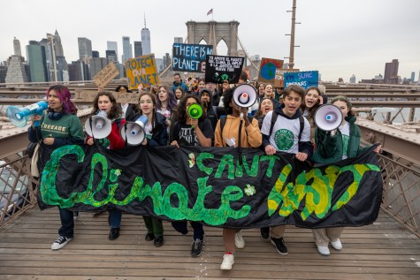 A line of activists with megaphones and signs stand in front of a large crowd of people. They hold a black banner with green text which reads “Climate Can’t Wait.” They are marching across the Brooklyn Bridge. The New York City skyline is in the background.
