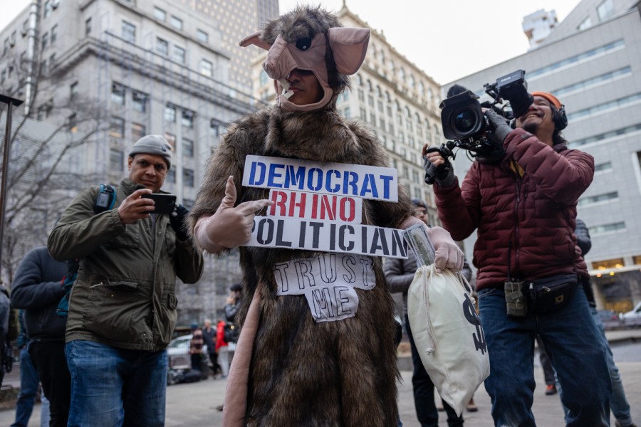 A rally attendee dressed in a gray mouse costume holding signs that read Democrat Rhino Politician and Trust me demonstrates in Collective Pond Park while two media personnel record the attendee from behind.