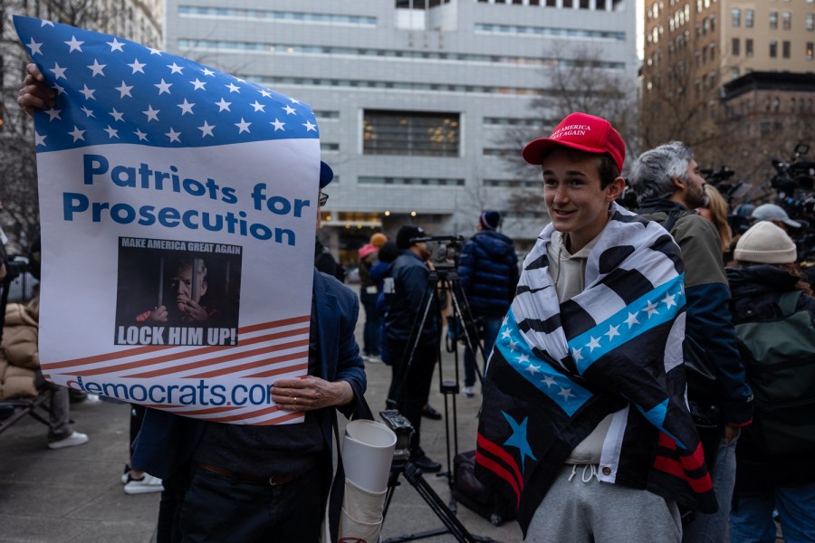 A boy wearing a red “Make America Great Again” cap and a republican flag wrapped around him standing next to man holding up a sign that reads, “Patriots for Prosecution” and has a photo of Donald Trump behind jail bars with the text, “MAKE AMERICA GREAT AGAIN, LOCK HIM UP!” and the website “democrats.com” written on it.