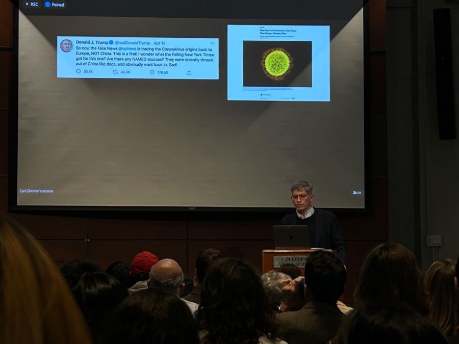Doctor Carl Zimmer speaks from the wooden podium in Hemmerdinger Hall. An audience sits in front of him. Behind him is a projector screen displaying two images.