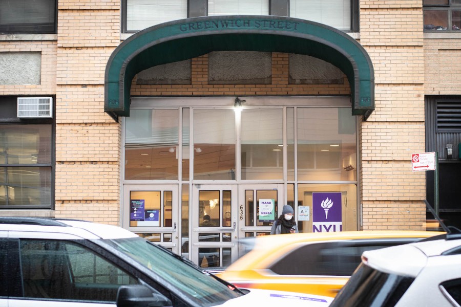 The entrance to N.Y.U’s Greenwich residence hall. Many cars pass by on the street. There is a large N.Y.U logo on the glass door.