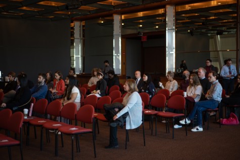 A room with people sitting on red chairs. Some chairs are empty with a notecard and pencil on top.