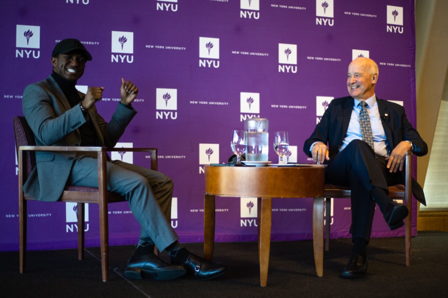 Ron Hall, a man in a gray suit and Andrew Hamilton, a man in a black suit, sit on a platform with a purple backdrop with the N.Y.U. logo.