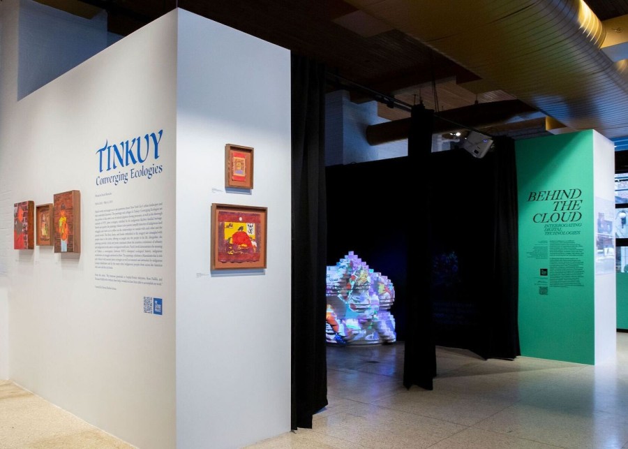 A wide gallery space, with a white wall on the left side of the room that has blue-colored text and pieces of art in brown frames. Behind it are black curtains and a large multicolored sculpture made of thin layers. On the right side of the image there is a green wall with black text.