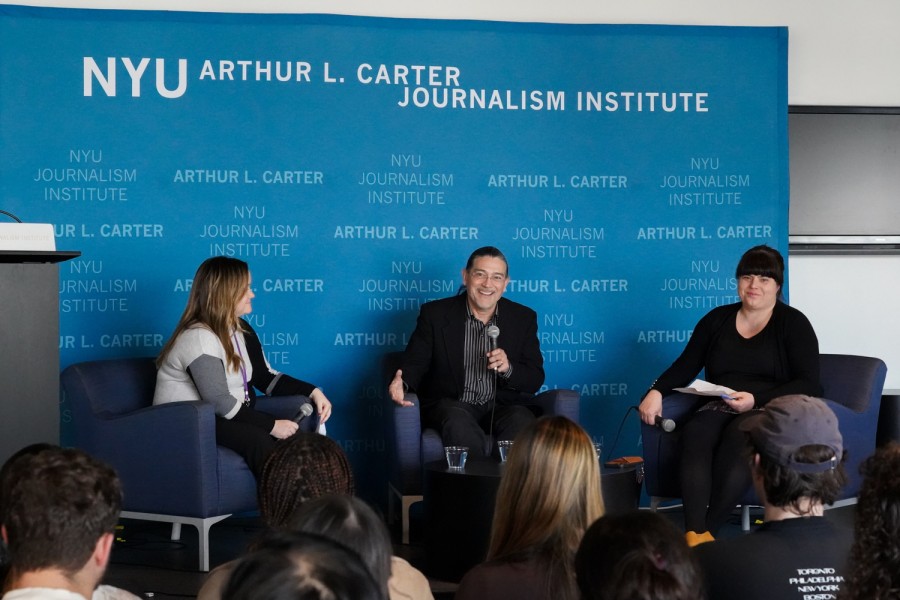 Robert Santos, the U.S. Census Bureau director, sits on a stage with two hosts on each side of him, speaking to the audience with a microphone in his hand. The backdrop behind him indicates that the event is at N.Y.U.’s Arthur L. Carter Journalism Institute.