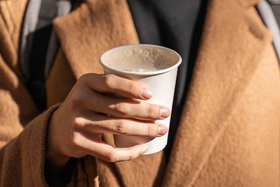 A hand holds a cup of coffee with foam on top. The cup is white and the person holding the coffee is wearing a beige coat.