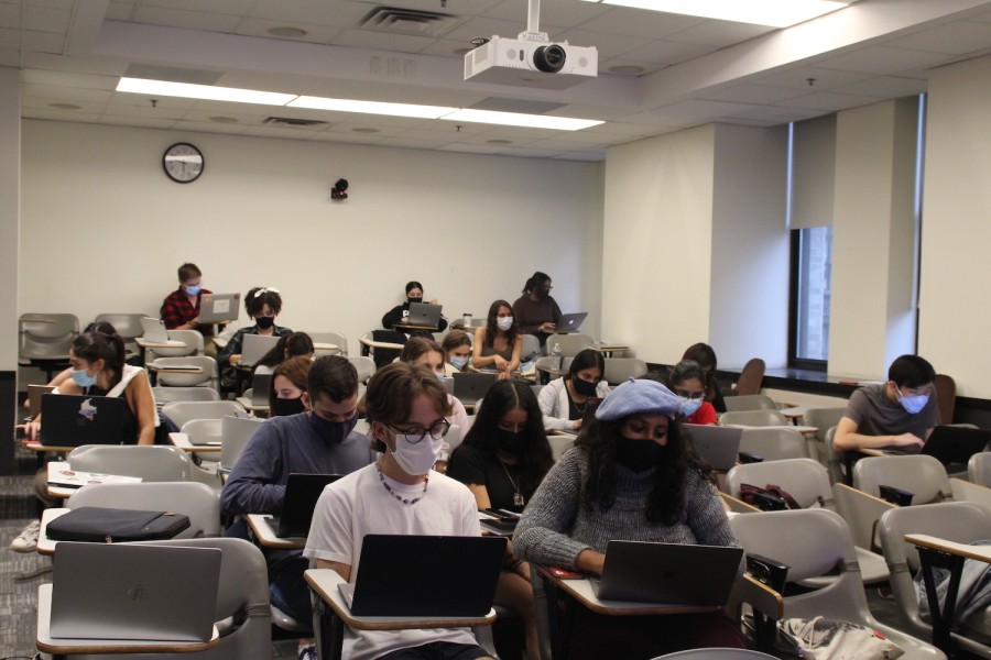 A classroom filled with students, who are wearing masks, working on their laptops. An analog clock hangs on the wall behind them.