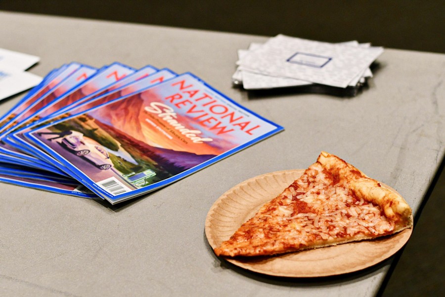A slice of pizza in a paper plate on a table next to a stack of magazines.  The name of the magazines is as follows "NATIONAL REVIEW."