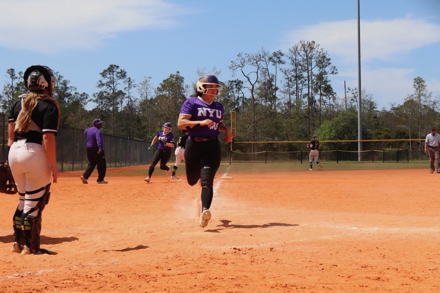 A softball player runs across a field while wearing a helmet and a purple jersey with the text “N.Y.U.” on it.