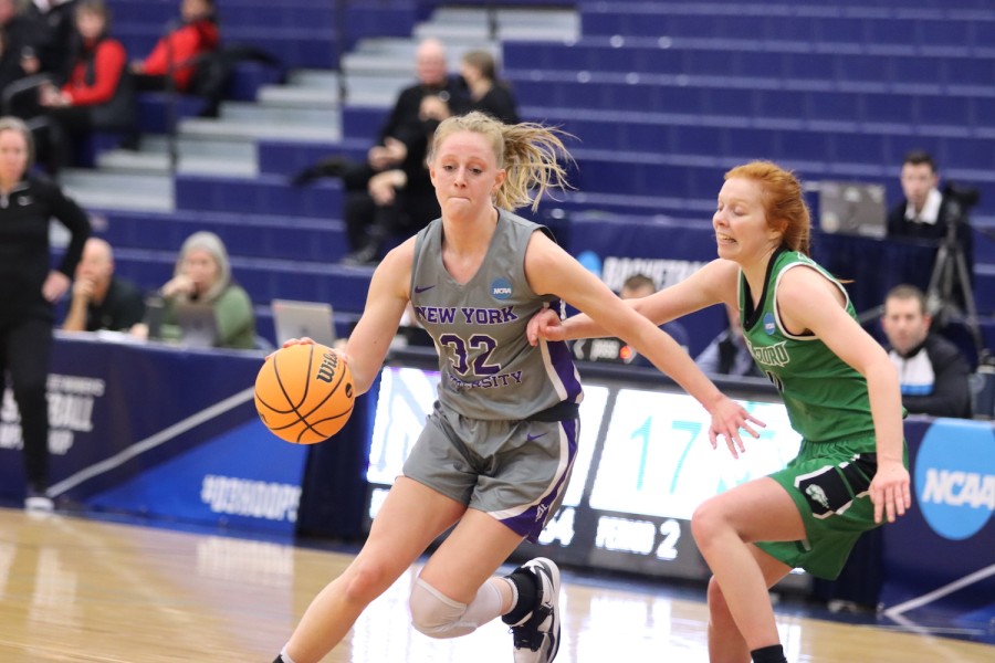 Photograph of an N.Y.U. basketball player with blond hair dribbling a basketball on a court. She is standing next to another player, who wears a green uniform.