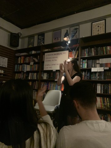 In a dimly lit room filled with shelves of books, a speaker wearing a black shirt speaks to an audience.