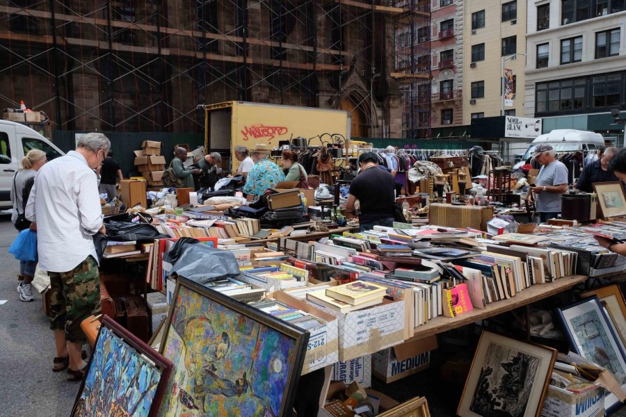 A flea market with people browsing in between the stalls. There are many books placed on the tables and paintings placed on the ground.