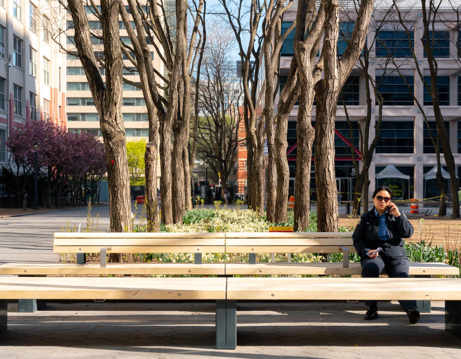 An FDNY employee is making a call while sitting on one of the new park benches. The three benches are long and feature metal frames with unfinished wood surfaces. A person sits on the right side of one bench.