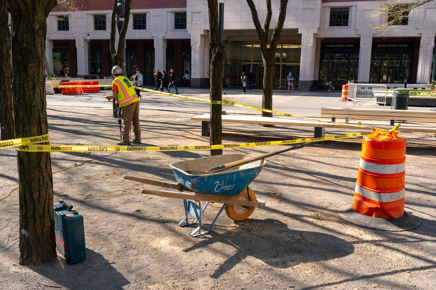 Construction work is being done in MetroTech’s park. There is a blue wheelbarrow (center), an orange construction item (right), and a construction worker in a reflective jacket (left) appearing to be working.