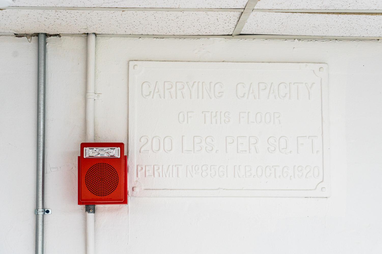 A sign reading "carrying capacity of this floor two hundred pounds per square foot" painted over in white on the wall of Rogers Hall's interior, bordering the ceiling. A red fire alarm is hung next to the sign.