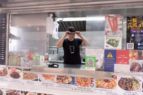 Mrs. Wang stands inside of her truck, wearing a black shirt and adjusting her hat. The outside of the food truck displays the menu.