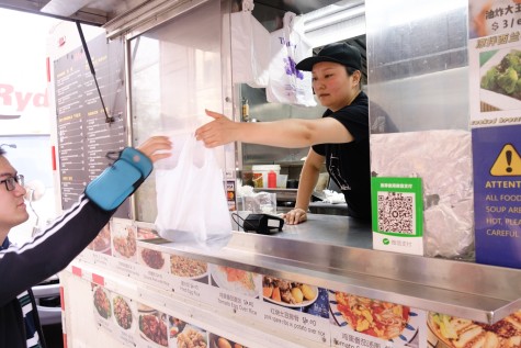 Mrs. Wang is wearing a black shirt as she hands a customer their food in a white plastic bag. The outside of the food truck displays the menu.