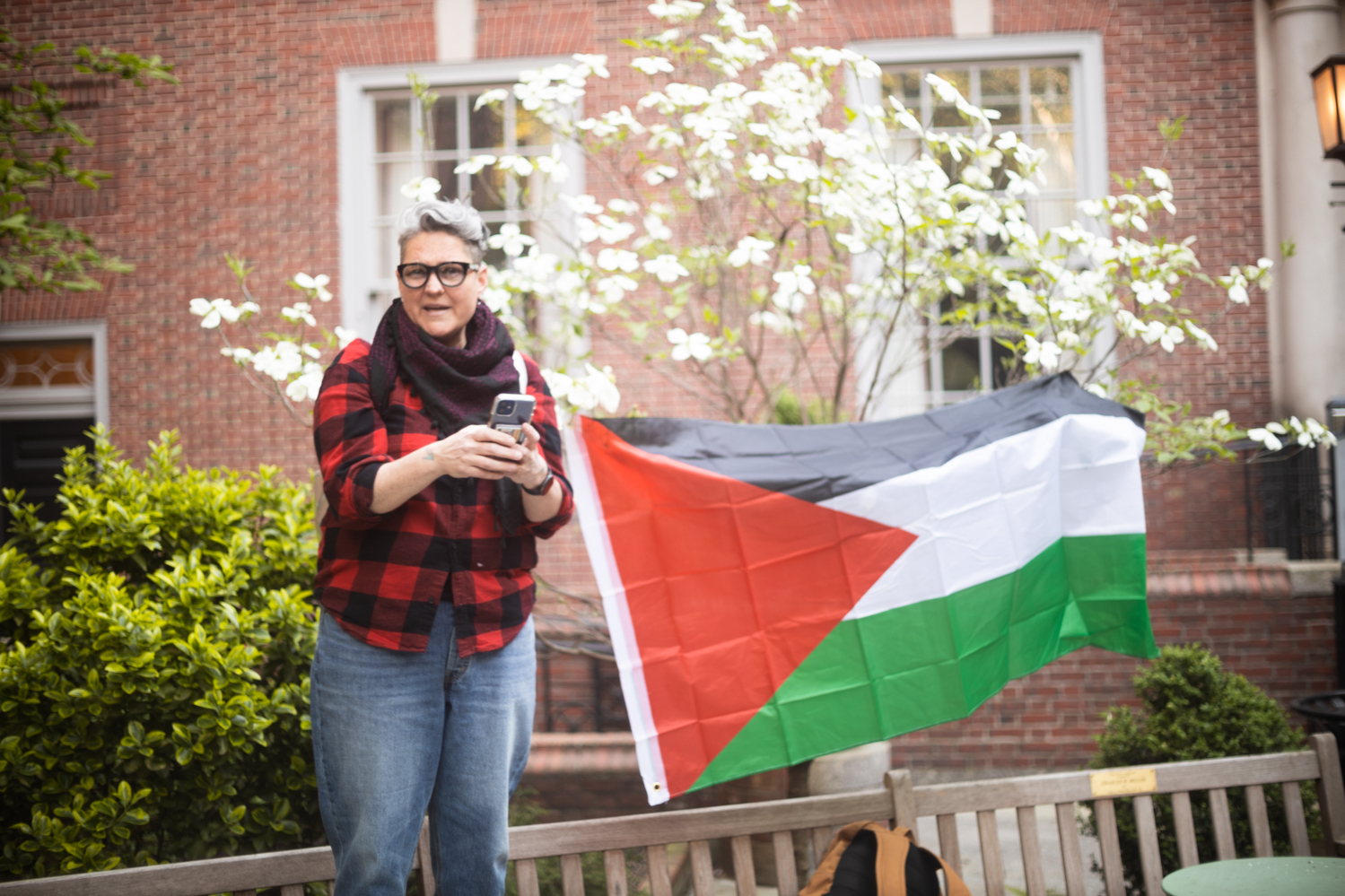 Emmaia Gelman, dressed in a red and black plaid flannel and blue jeans, speaks in front of a Palestinian flag.