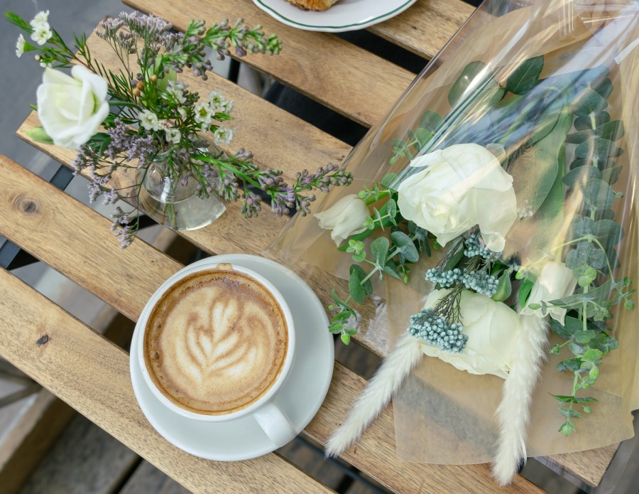 A cup of coffee and a bouquet of flowers placed on a wooden table outdoors.