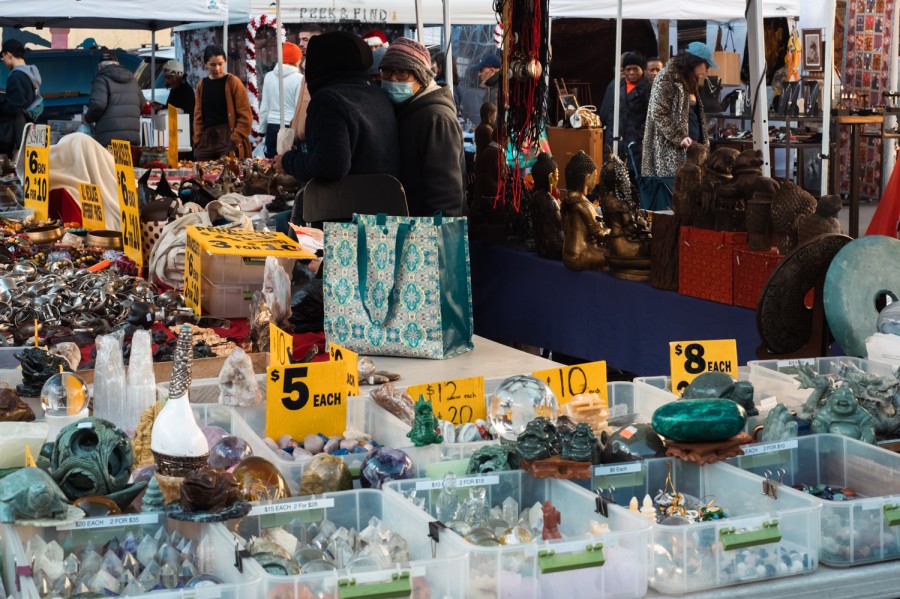 A stall at a flea market with many boxes holding different types of stones and crystals on a table. There are yellow price tags next to some of the boxes. People are browsing the stall in the back.