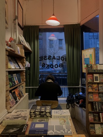 A white room filled with bookshelves on all sides. A light with a red cover hangs from above. In the background is a window with the inverted text “Molasses Books” printed on it.