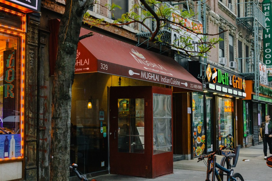 The façade of a restaurant with red frames and awning. Its exterior awning reads “ Mughlai Indian Cuisine.”