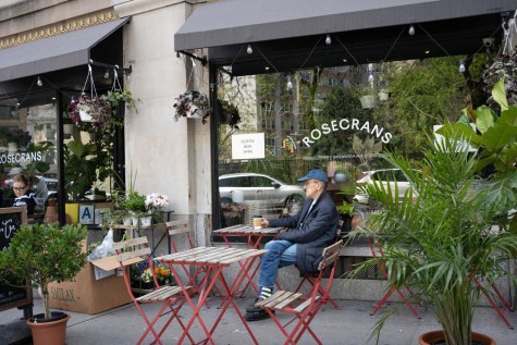A man wearing a blue cap sits on a chair outside of a cafe, and there are potted plants and flowers placed outside the windows