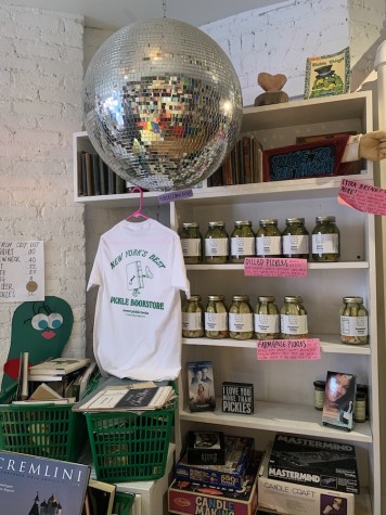 A book titled “Cremlini” in the foreground and behind, two green baskets filled with books. A white shirt with green text that reads “New York’s Best Pickle Bookstore” hangs from a shelf, on which are jars of pickles.