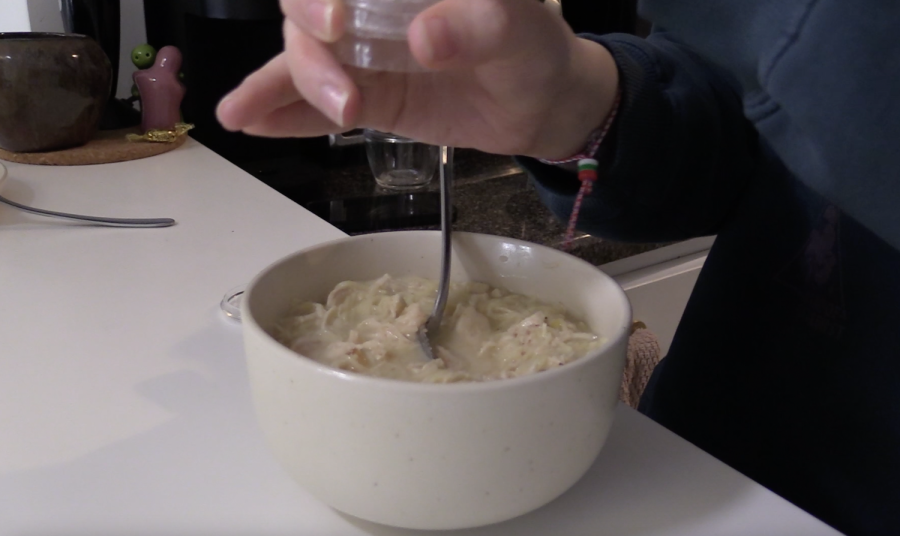 A hand holding a bottle above a bowl of noodles that is placed on a kitchen counter.