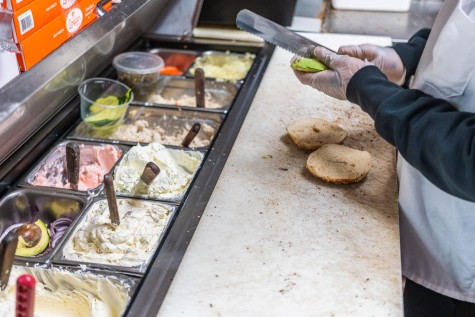 An employee of Bagel Bob’s slices an avocado while preparing a bagel. There are various cream cheese flavors in front of the employee.