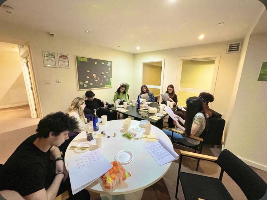 Eight people sit around a circular white table and a square black table, which are placed next to each other. They are reading from sheets of paper, with various cups and foods on the tables for seder. They are in a white-walled room with two glass doors and small framed pictures displayed on the walls.