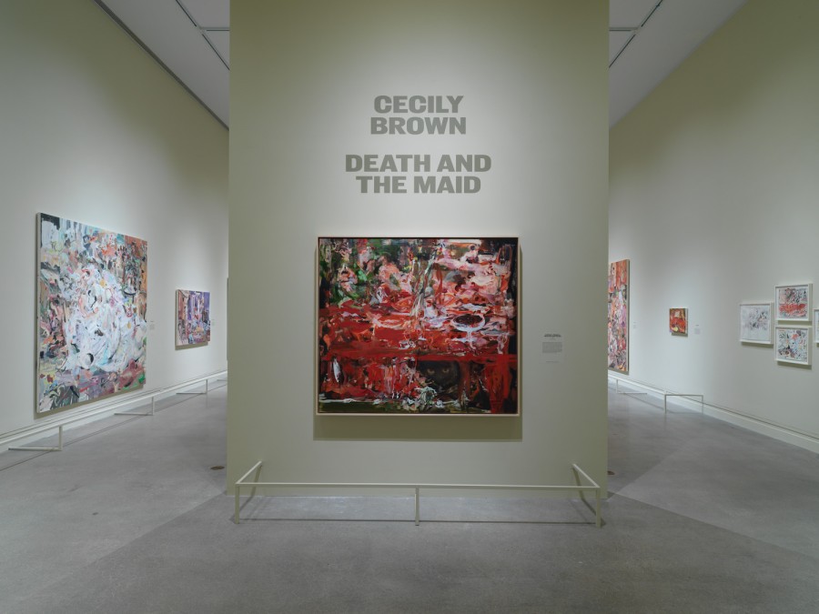 A red, brown and white painting hanging on a cream colored wall inside the Metropolitan Museum of Art. There is a cream colored railing around the base of the wall, and “Cecily Brown, Death and the Maid” is written in capitalized gray letters above the painting.