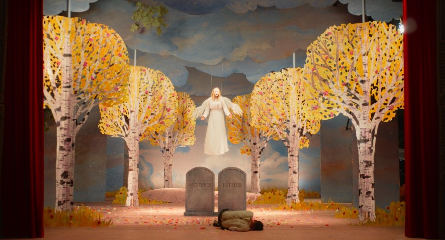 A woman with blonde hair wearing a white dress with wings while being held up by strings above two gravestones bearing the words “MOTHER” and “FATHER” on a stage with opened red curtains. Curled in a ball in front of the gravestones lies a man in a tan shirt and brown pants.z