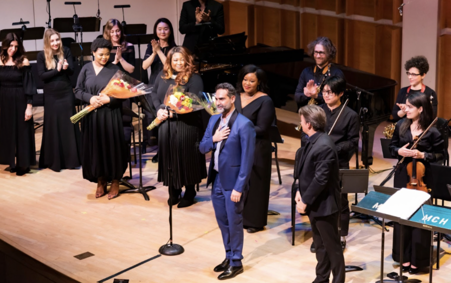 A group of people wearing black formalwear stands on an opera stage holding bouquets of flowers and clapping around a man wearing a blue suit with his hand over his heart looking up at the audience.