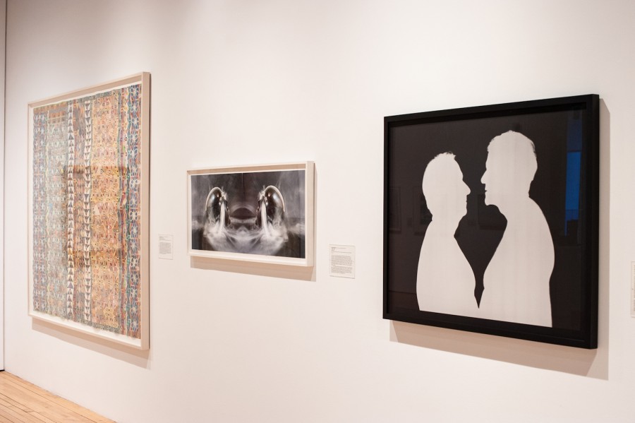 A white wall with three pieces of art displayed on it. On the left, a large textile artwork. In the middle, a small abstract black-and-white print. On the right, a slightly larger black-and-white print of silhouettes of two bodies facing each other.