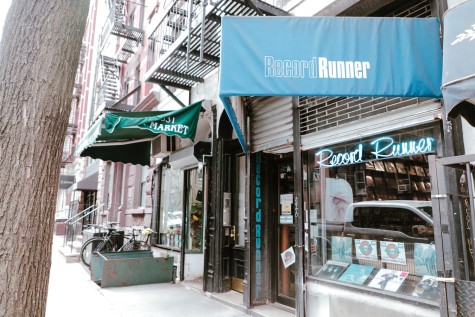 The Record Runner store. Above the store is a blue awning which has “Record Runner” printed on it. Behind the window of the store is a blue neon sign that reads “Record Runner.”