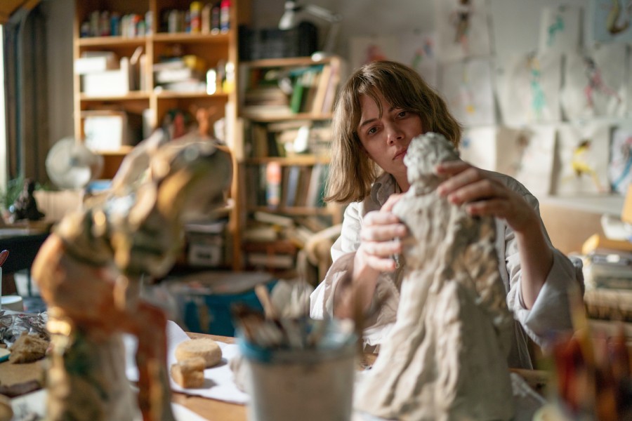 Actress Michelle Williams as the character Lizzy in the film “Showing Up.” She is working on a sculpture inside a studio with a bookshelf in the background.
