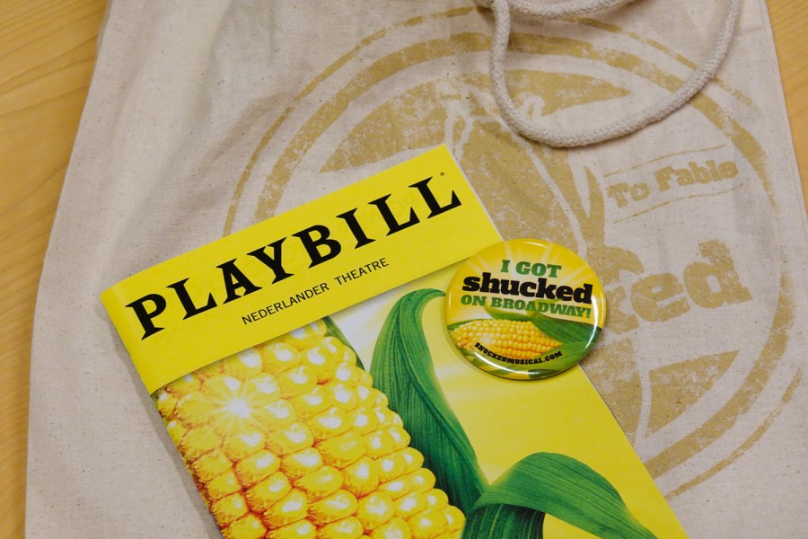 A+Playbill+with+an+image+of+corn+on+the+cover+is+placed+on+top+of+a+fabric+bag.+A+button+is+pictured%2C+with+an+image+of+corn+and+the+text%2C+%E2%80%9CI+got+shucked+on+Broadway%21%E2%80%9D