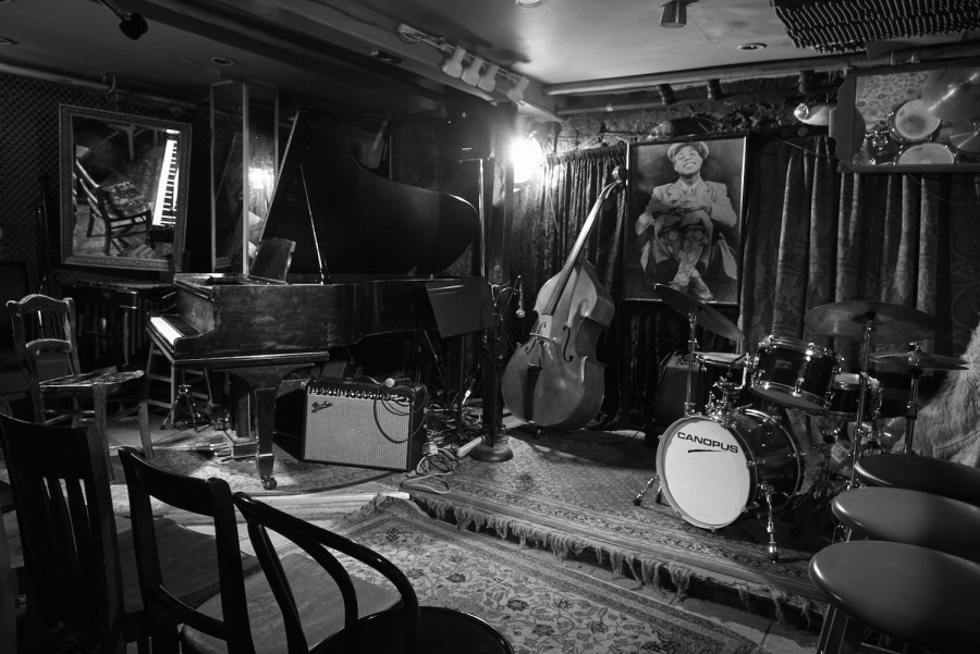 A black-and-white photo of a stage with two rugs, a grand piano, a double bass, an amp and a drum set.