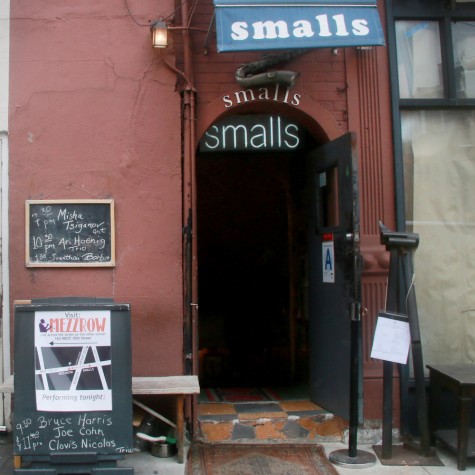 The exterior of a jazz club. A blue awning hanging above a black door in the middle of a red facade reads “Smalls.”