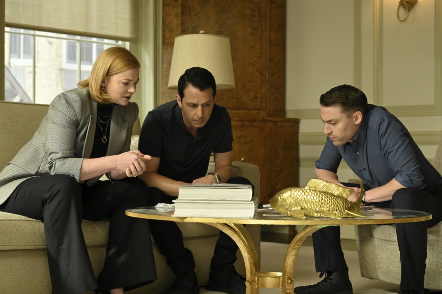A woman with short blonde hair wearing a gray blazer and black pants, a man with short black hair wearing a black collared shirt and black pants, and a man with short black hair wearing a navy button-up and black pants all sit on a couch looking at a coffee table that has a stack of documents on it.