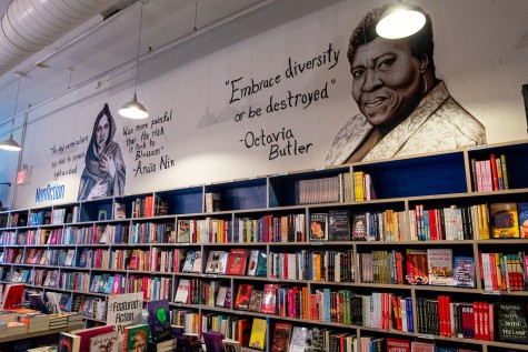 The interior of Bluestockings Cooperative. At the bottom is a row of shelves stacked with books. Above them is a mural that depicts Octavia Butler with a quote that reads “Embrace diversity or be destroyed.”