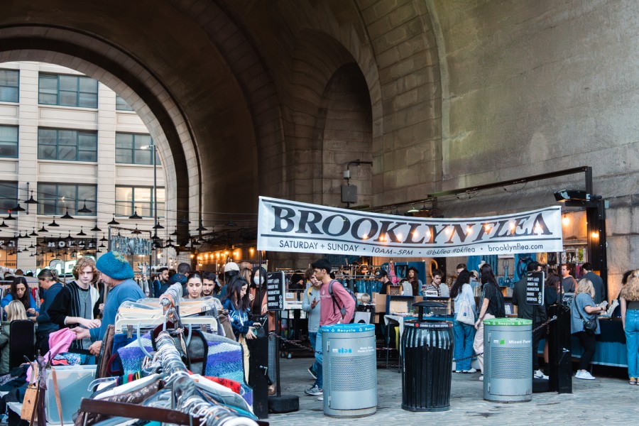 A flea market under the Manhattan Bridge with a white banner that reads “BROOKLYN FLEA.” There are people browsing in between the stalls.