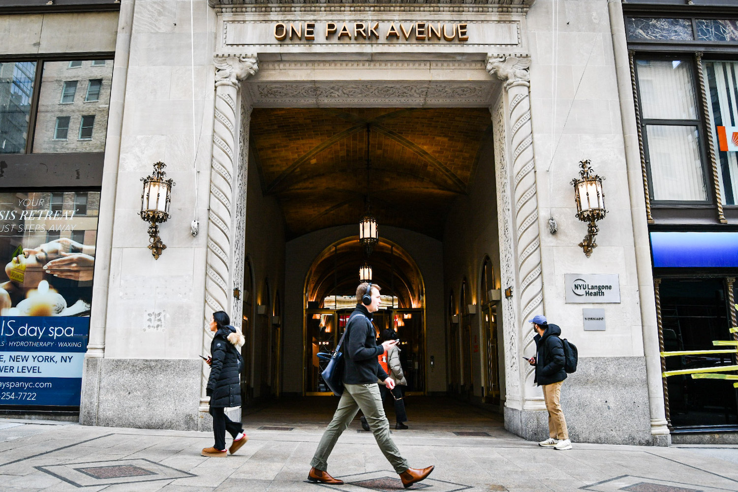 A man wearing headphones walks in front of the exterior of One Park Avenue. “N.Y.U. Langone Health” is written on a plaque outside.