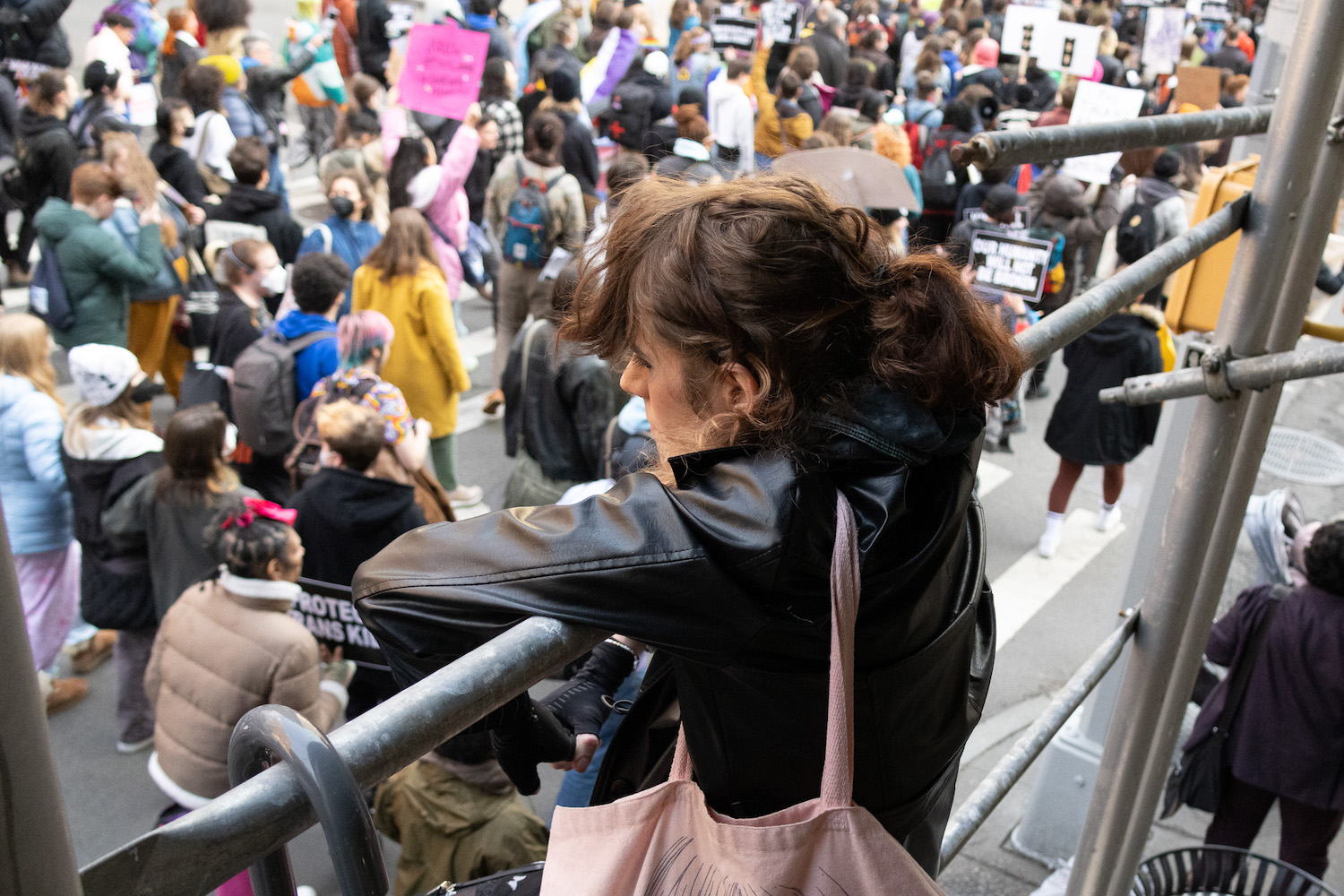 A person seen from above and behind, sits on construction scaffolding overlooking protesters marching down a street. Her cane hangs from the scaffolding beside her.